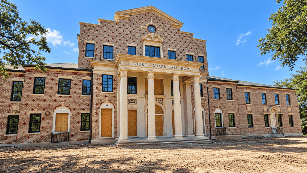 Exterior view of the Touro-Shakespeare Home, a brick building with columns and boarded windows, set against a blue sky.
